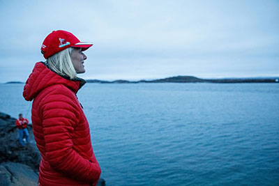 Catherine McKenna is wearing a bright red jacket & baseball cap. She is looking out on the ocean.
